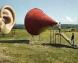 A surreal landscape by Erik Johansson photography. A giant ear stands next to a red megaphone, with people climbing stairs towards it.