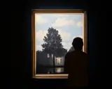 A person gazes at Magritte's "Empire of Light," a surreal scene of a house illuminated by a streetlamp under a daytime sky.