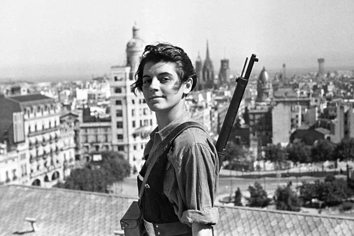 A young woman with a rifle slung over her shoulder stands on a rooftop, overlooking a city skyline. This is one of the most famous photographs of the Spanish Civil War.
