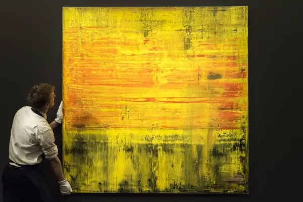 A man in white gloves carefully handles a large abstract painting with vibrant yellow and orange hues, a testament to Gerhard Richter's net worth.