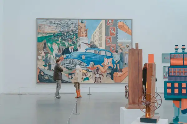 Two people observe a large, colorful mural depicting a chaotic scene with a blue car, factories, and people, offering a glimpse into the art market definition of diverse artistic expression.