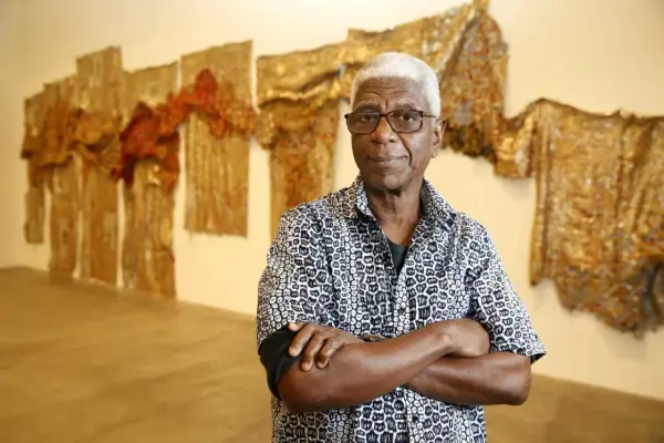 A distinguished man with white hair and glasses, arms crossed, stands before a wall adorned with shimmering, textured artworks, a testament to the enduring legacy of famous artists today.